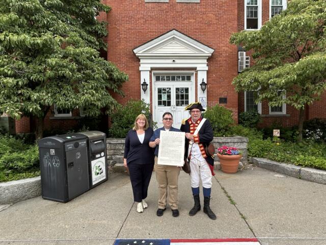 Three people stand outside a brick building holding a large document, with a costumed colonial-era figure on the right.