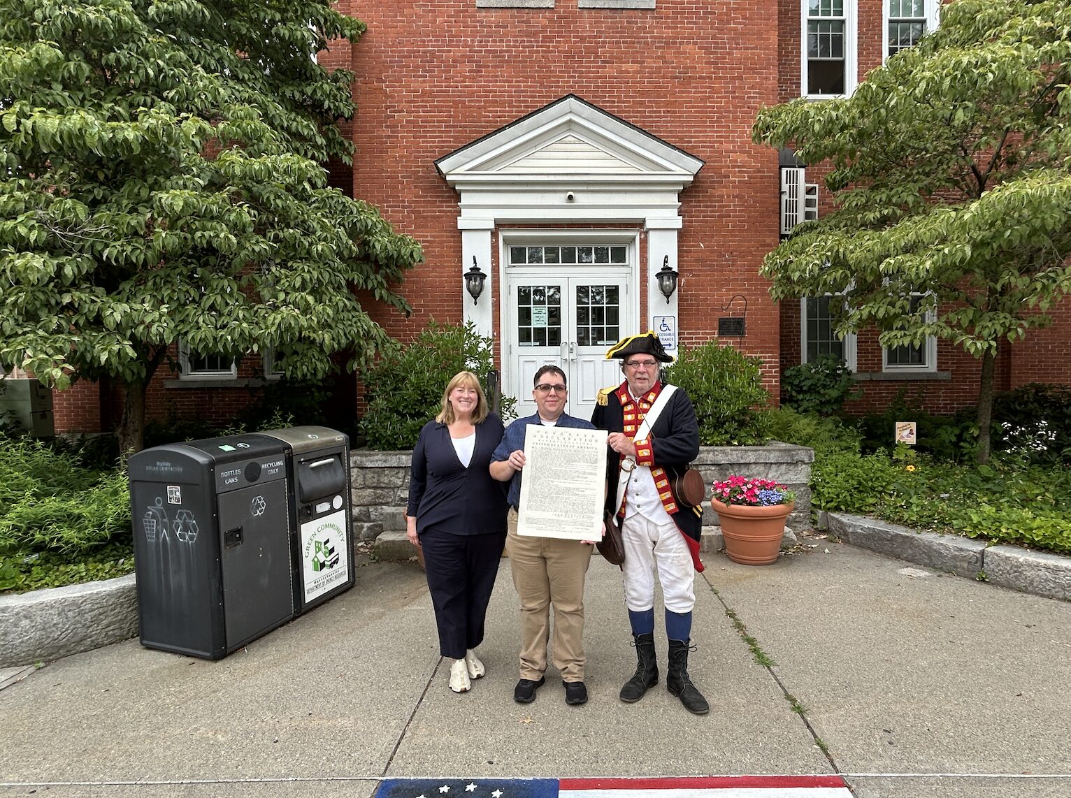 Three people stand outside a brick building holding a large document, with a costumed colonial-era figure on the right.