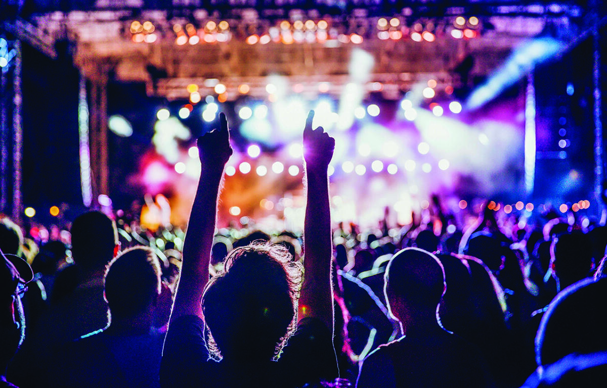 Crowd at a live concert with hands raised toward a brightly lit stage, colorful spotlights.