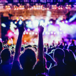 Crowd at a live concert with hands raised toward a brightly lit stage, colorful spotlights.