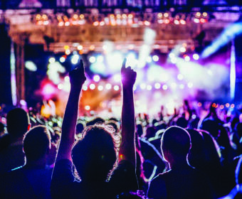 Crowd at a live concert with hands raised toward a brightly lit stage, colorful spotlights.