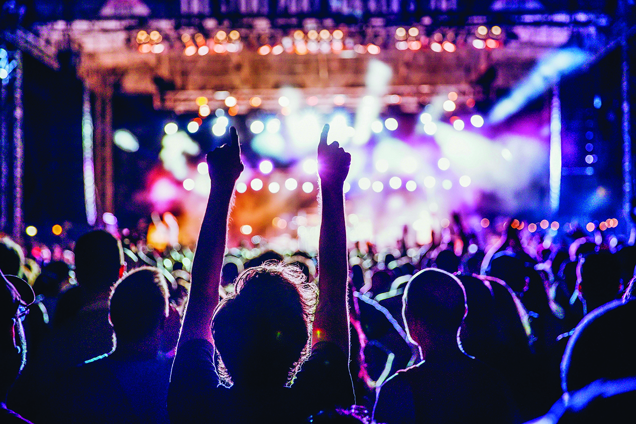 Crowd at a live concert with hands raised toward a brightly lit stage, colorful spotlights.