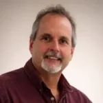 Portrait of a middle-aged man smiling, with short gray hair and a goatee, wearing a maroon shirt against a light background.